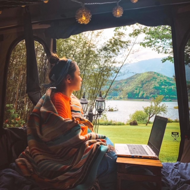 Girl camping sitting in roof tent with laptop looking at lake
