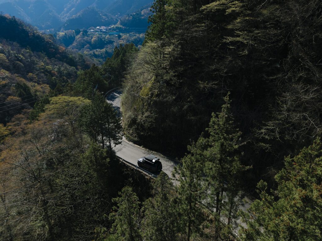 春の森を抜ける山道を走るLand Rover Defenderのキャンピングカーを上から見たところ