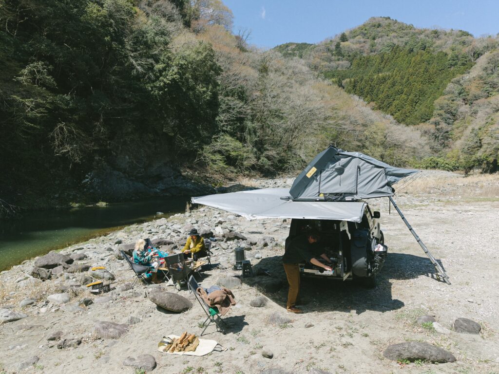 山あいの谷の河原のキャンプで、オーニングを広げたDefenderのキャンピングカー