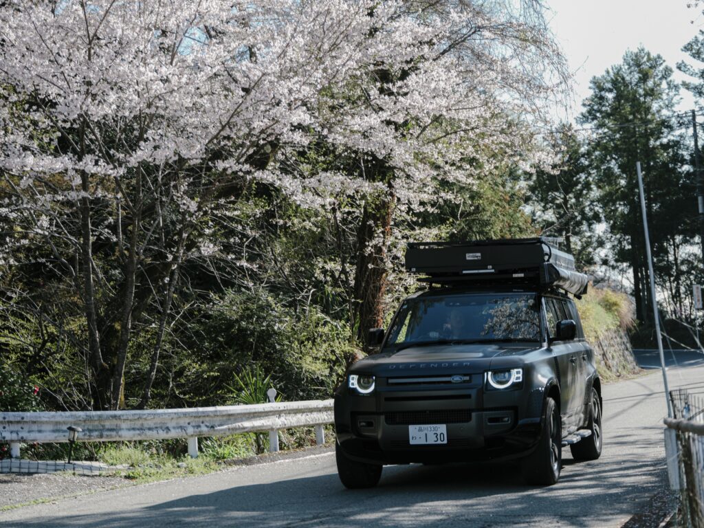 桜の下の山道を走るLand Rover Defenderのキャンピングカー