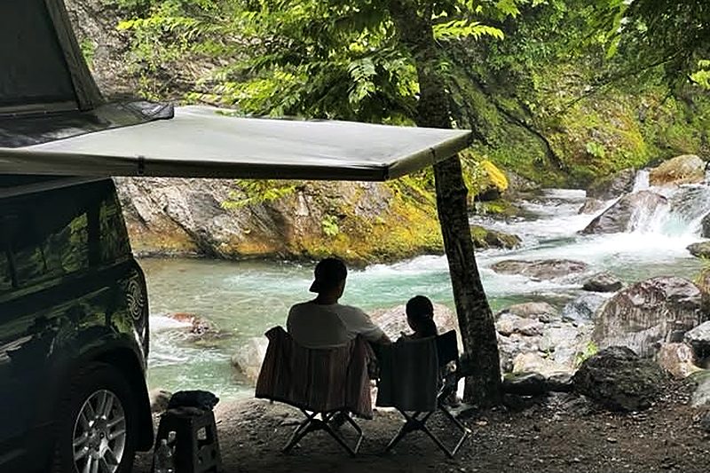 Two people sitting in camp chairs under a camper's awning, looking out over a clear mountain river at Owata Campground