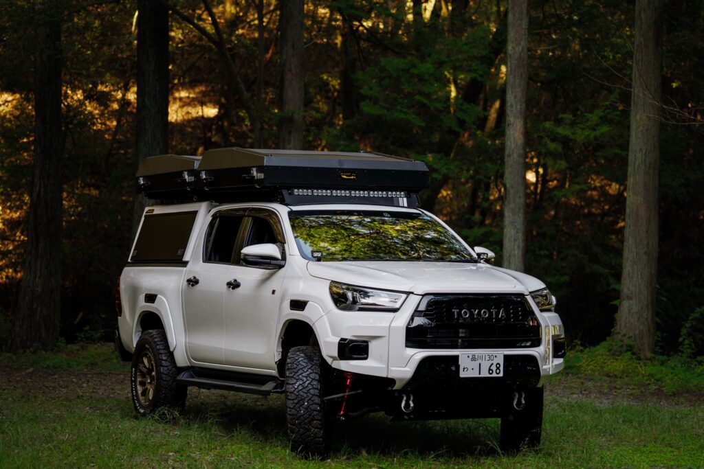 A white Toyota Hilux camper with its rooftop tent closed, parked in a cedar forest