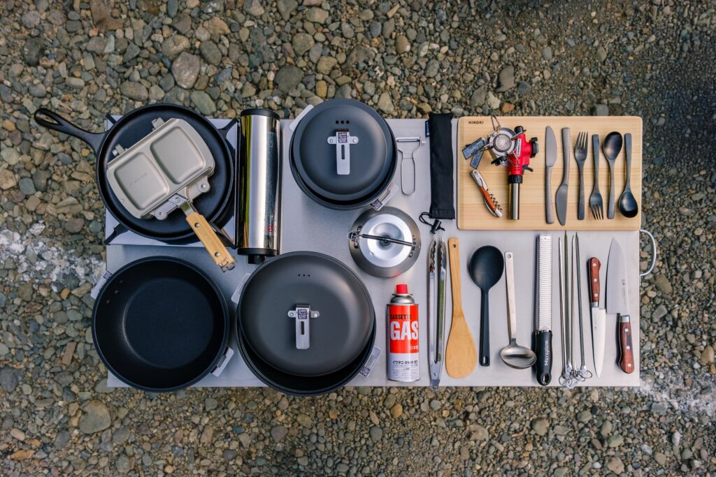 A camper's kitchen kit laid out on a board: pans, kettle, utensils, cups and gas
