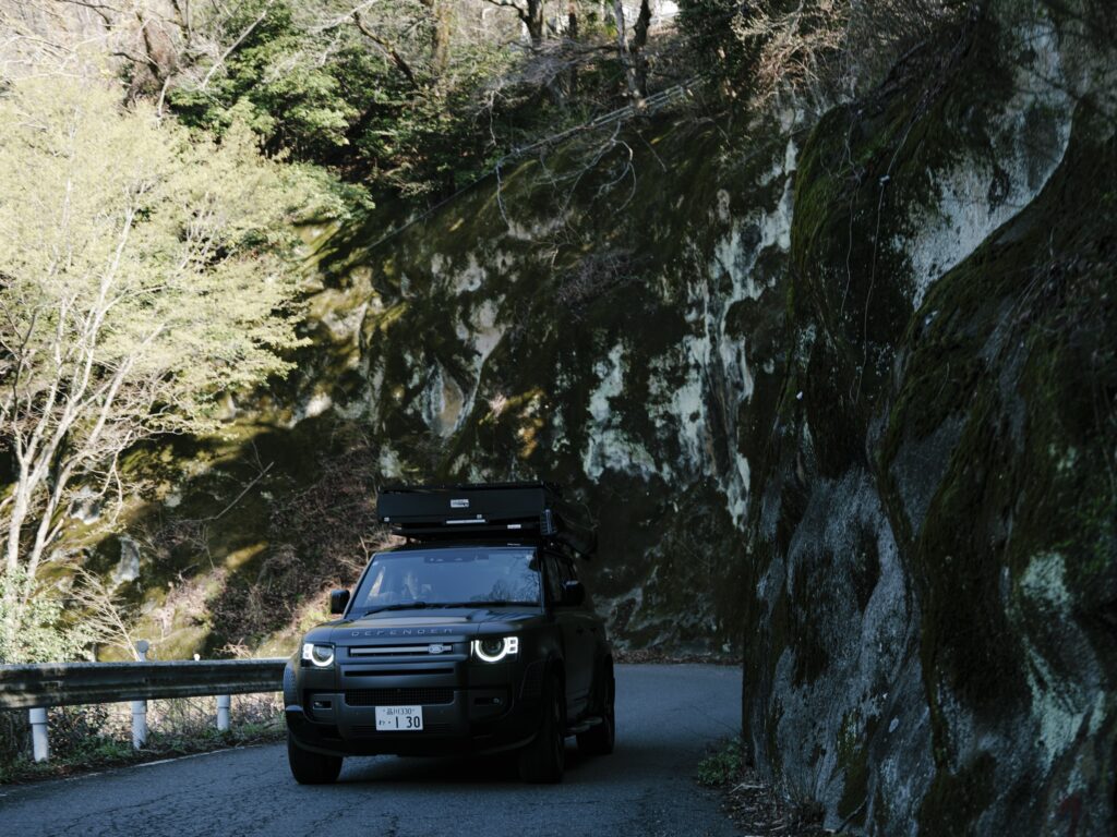 A Land Rover Defender camper on a narrow mountain road beneath a rock face