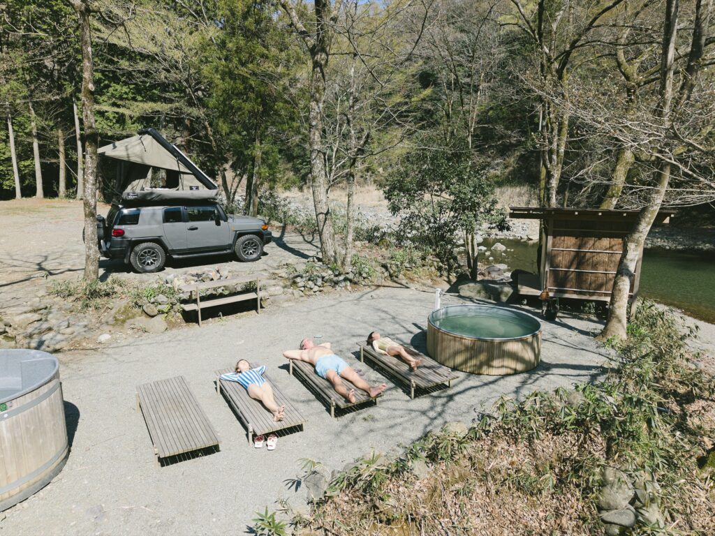 寝椅子と木の湯船がある川沿いのサイトに停まるキャンピングカーを、上から見たところ