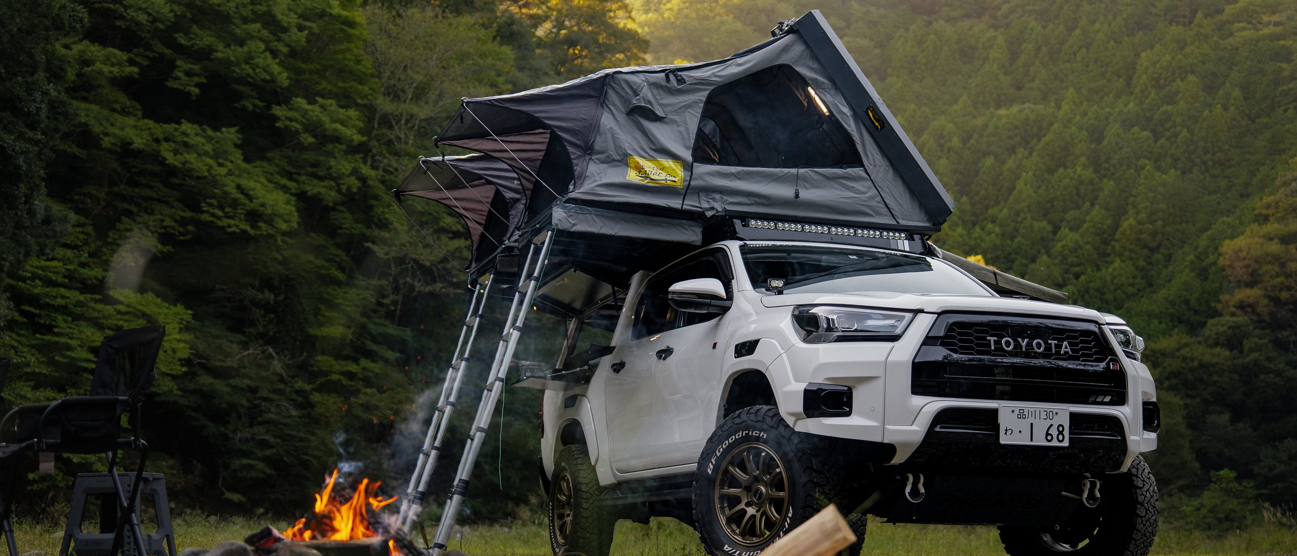 2つのルーフテントを広げてはしごを下ろしたHilux、手前に焚き火とキャンプチェア、背後に森の丘