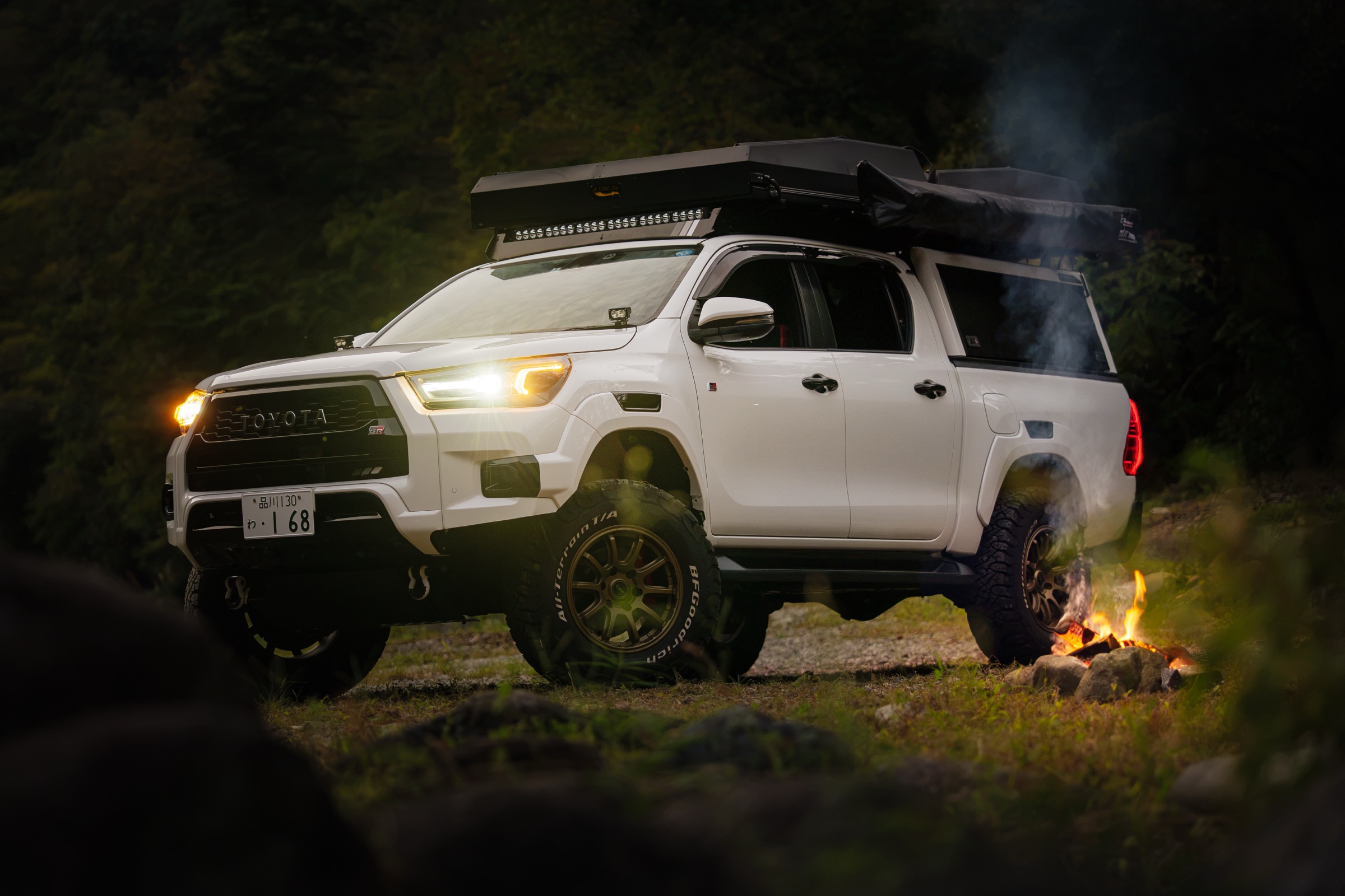 A white Hilux camper with its lights on beside a campfire at night