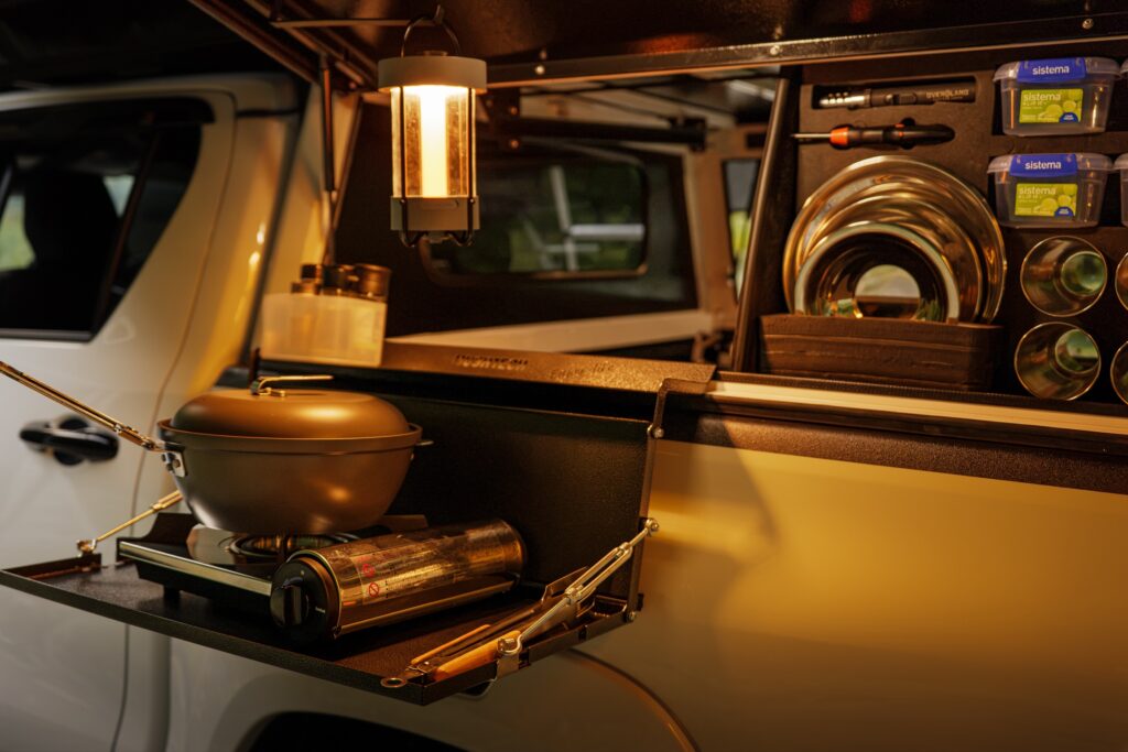 A camper's slide-out kitchen lit by a lantern, a gas cooker and kettle ready